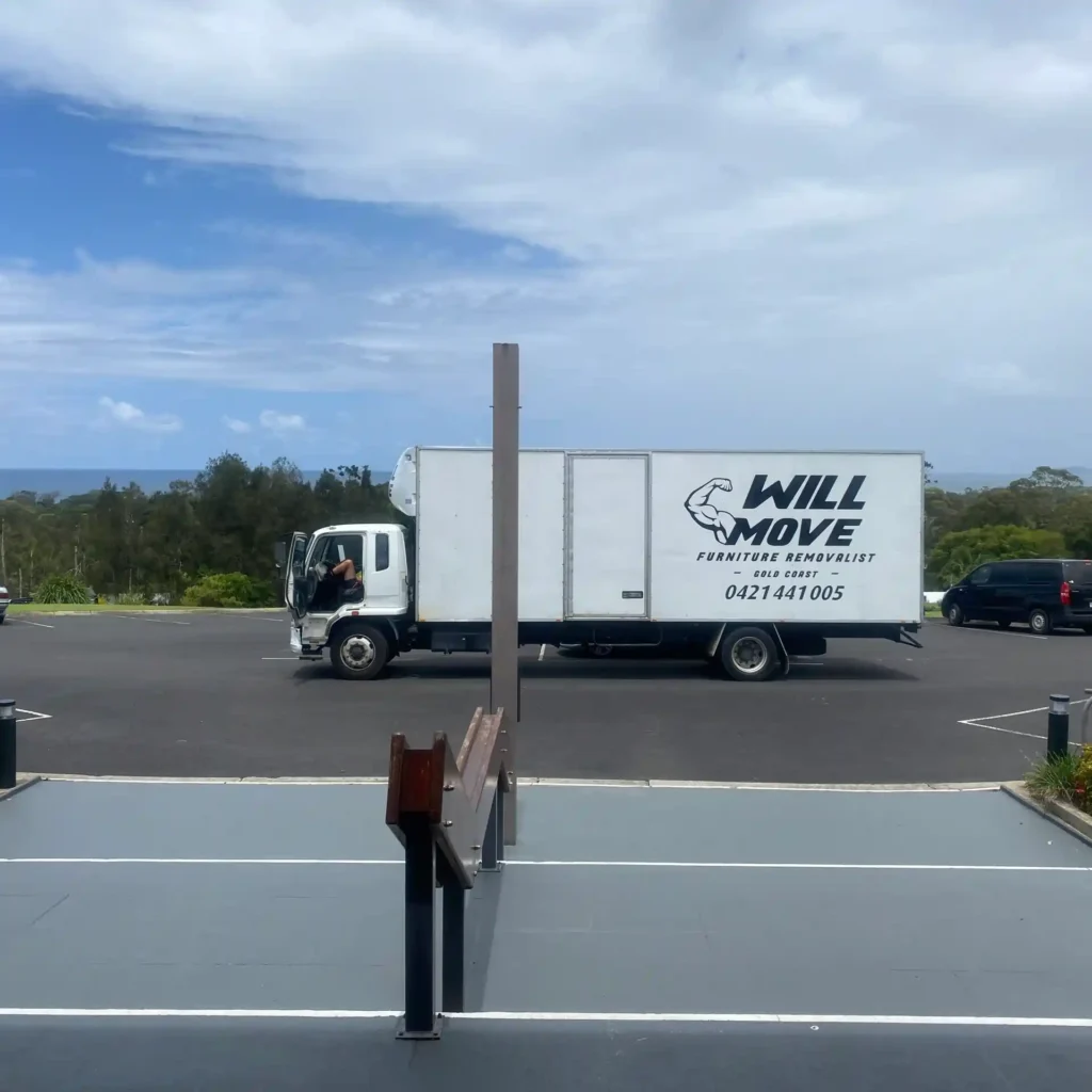 Image-21-scaled | Will Move Removalist White moving truck with 'Will Move' logo parked in a lot, ocean visible in the background under a blue sky.