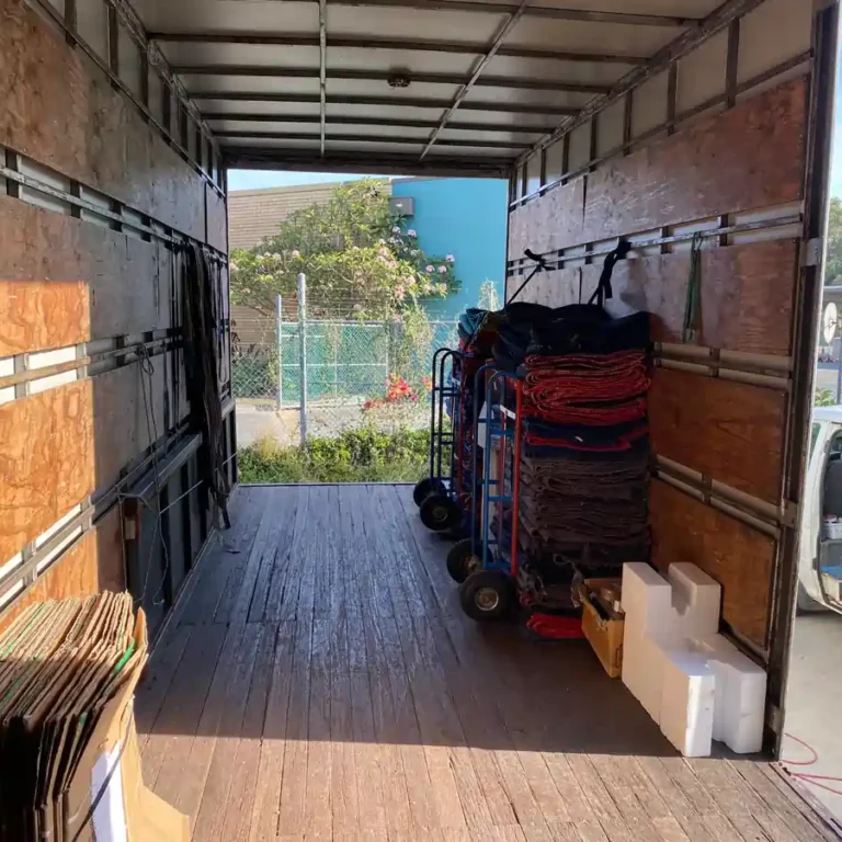 Interior of an empty moving truck with wooden side walls; red and gray mats stacked on rolling carts along the right side near the doorway.
