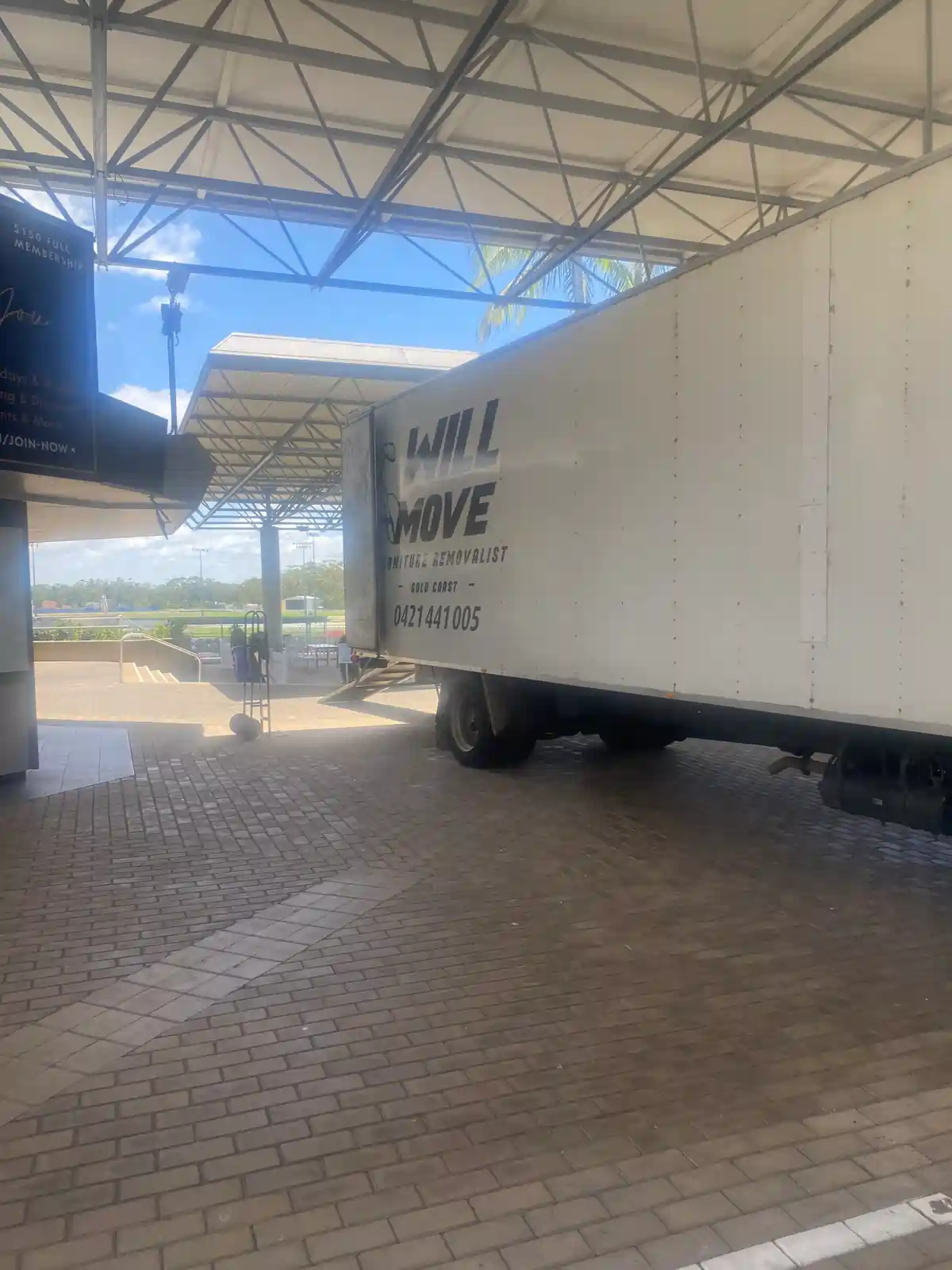 White moving truck trailer labeled 'WILL MOVE' parked under a metal canopy at a brick-paved loading area with blue sky.