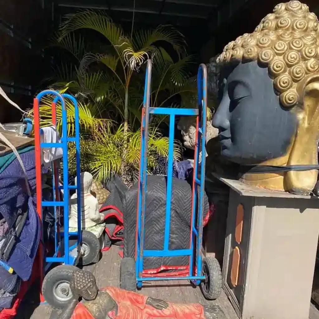 Industrial storage area with a large Buddha head statue on a pedestal beside blue and red moving carts and scattered statues outdoors.