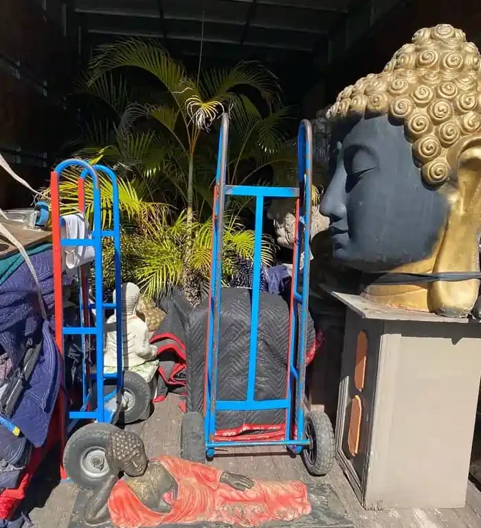 Local-Removals | Will Move Removalist Industrial storage area with a large Buddha head statue on a pedestal beside blue and red moving carts and scattered statues outdoors.
