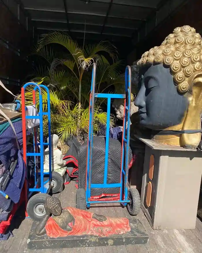 Local-Removals | Will Move Removalist Industrial storage area with a large Buddha head statue on a pedestal beside blue and red moving carts and scattered statues outdoors.