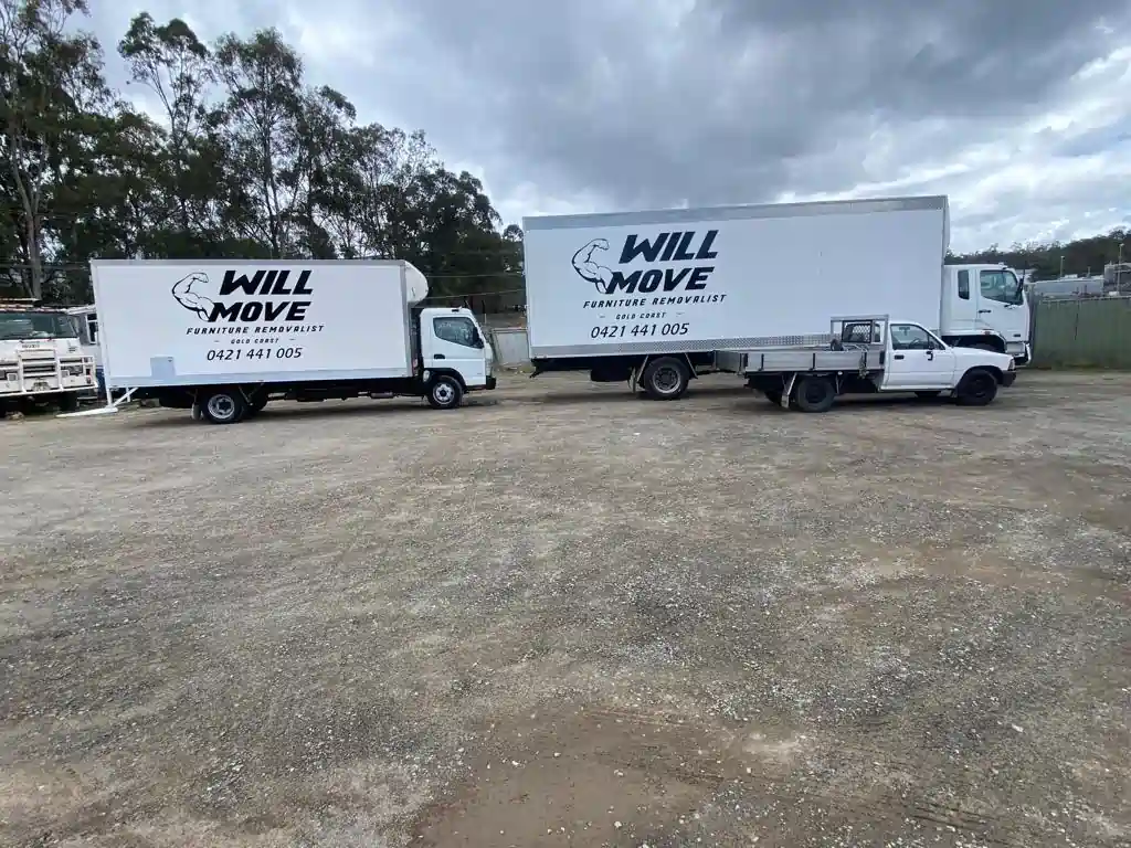 WIll-Move-Van-3 | Will Move Removalist Two white Will Move furniture removal trucks parked on a gravel lot beneath a cloudy sky.