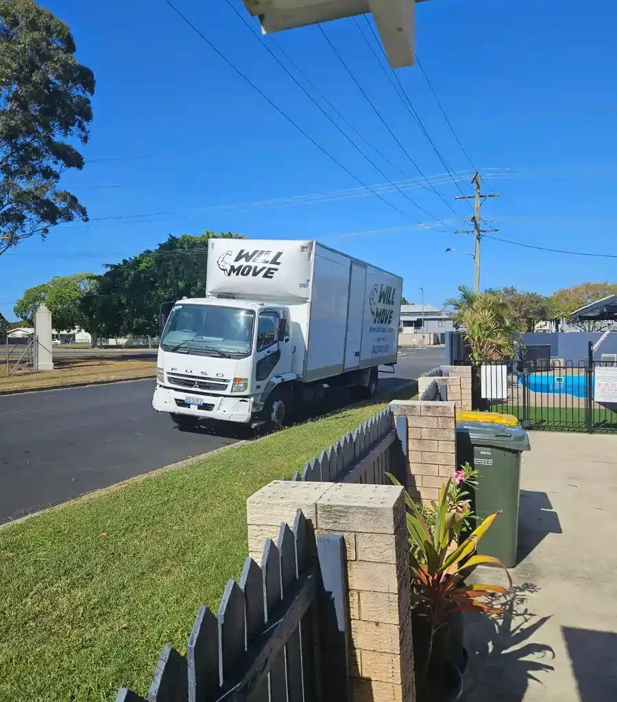 White moving truck labeled 'WILL MOVE' parked on a suburban street beside a grassy curb and fence.