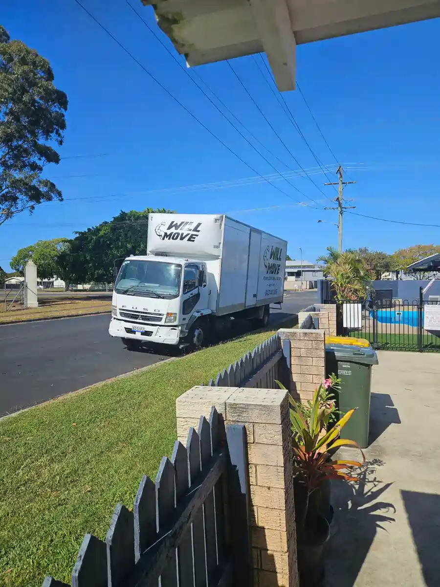 White moving truck labeled 'WILL MOVE' parked on a suburban street beside a grassy curb and fence.