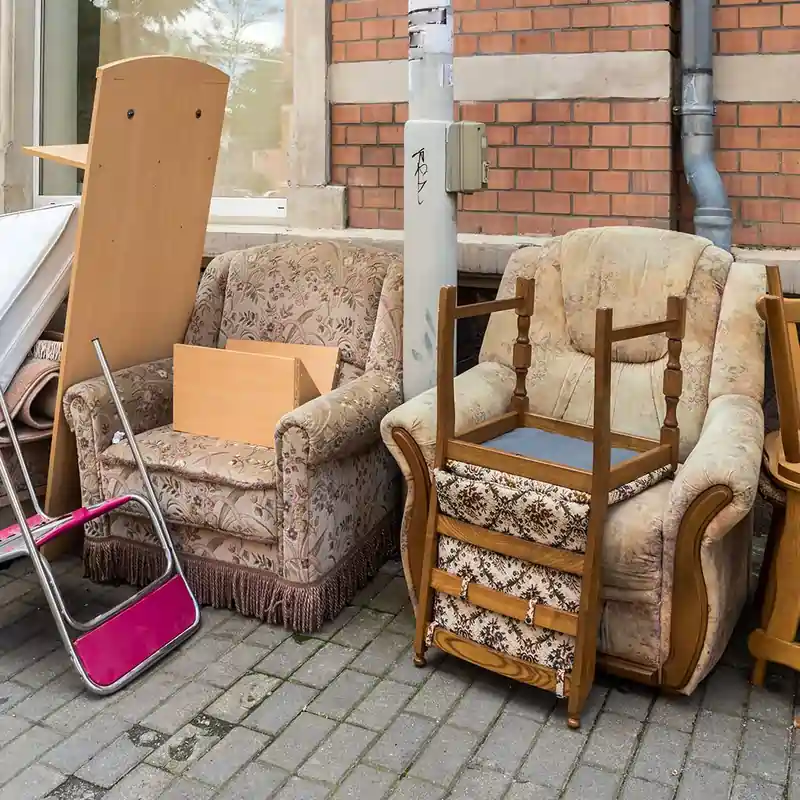 Discarded furniture stacked on a sidewalk: two floral armchairs and a wooden chair frame among other items.