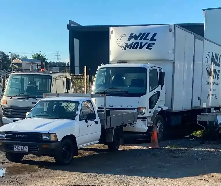 White Toyota pickup towing a flatbed behind a large Will Move moving truck in a yard with a cone nearby
