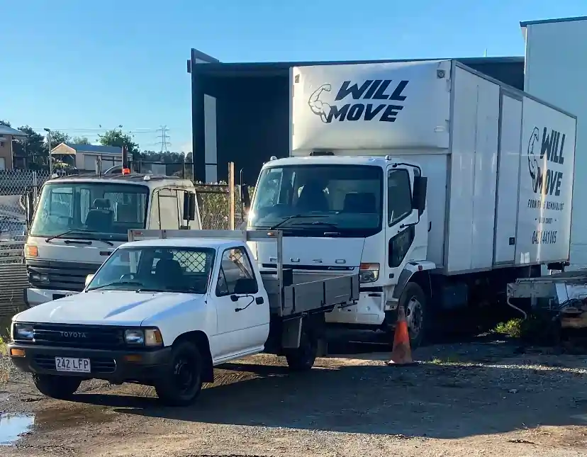 White Toyota pickup towing a flatbed behind a large Will Move moving truck in a yard with a cone nearby