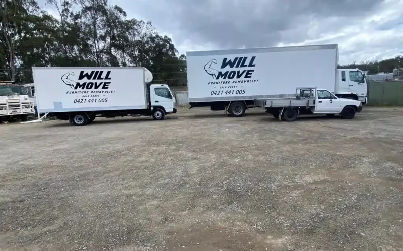 Two white Will Move furniture removal trucks parked on a gravel lot beneath a cloudy sky.