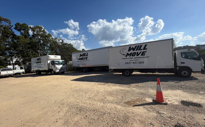 Three white Will Move moving trucks parked on a gravel lot under a bright blue sky with fluffy clouds and a red-orange traffic cone in the foreground.