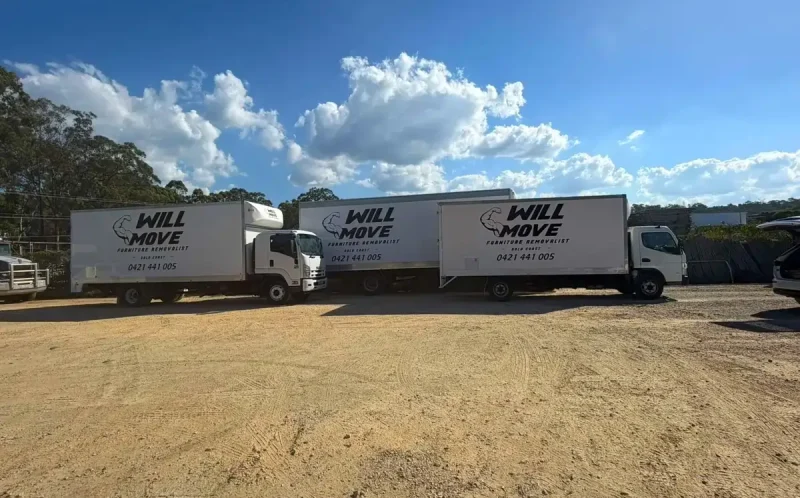 Three white Will Move moving trucks parked on a dirt lot under a sunny blue sky with clouds.